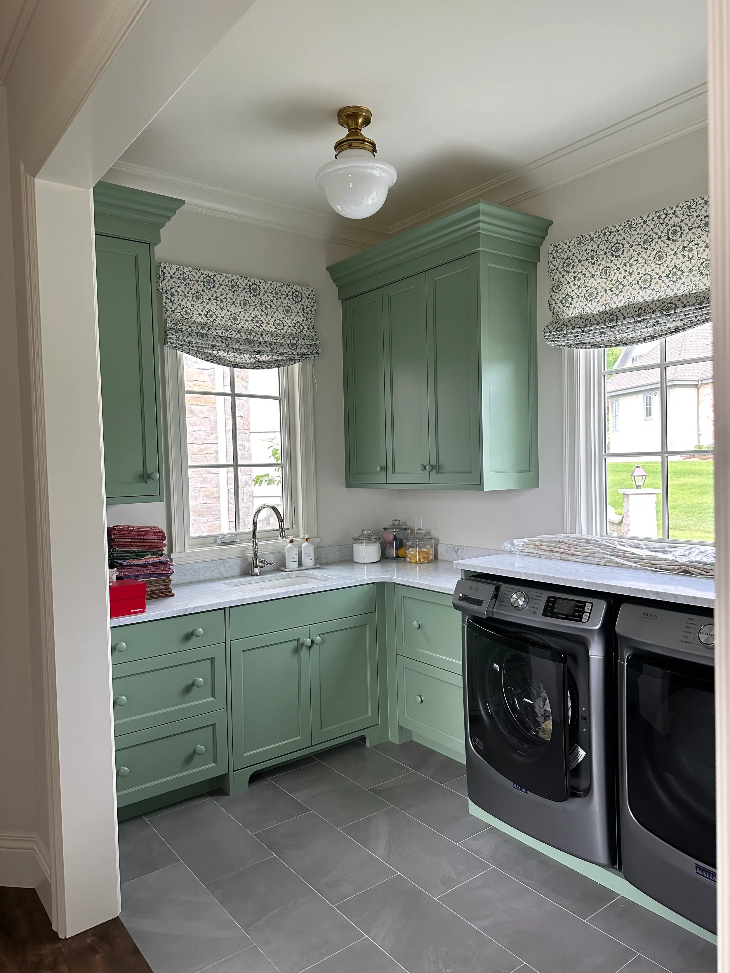 Sage green shaker laundry room cabinetry with marble countertop, farmhouse sink, silver gooseneck faucet, and patterned Roman window shades.
