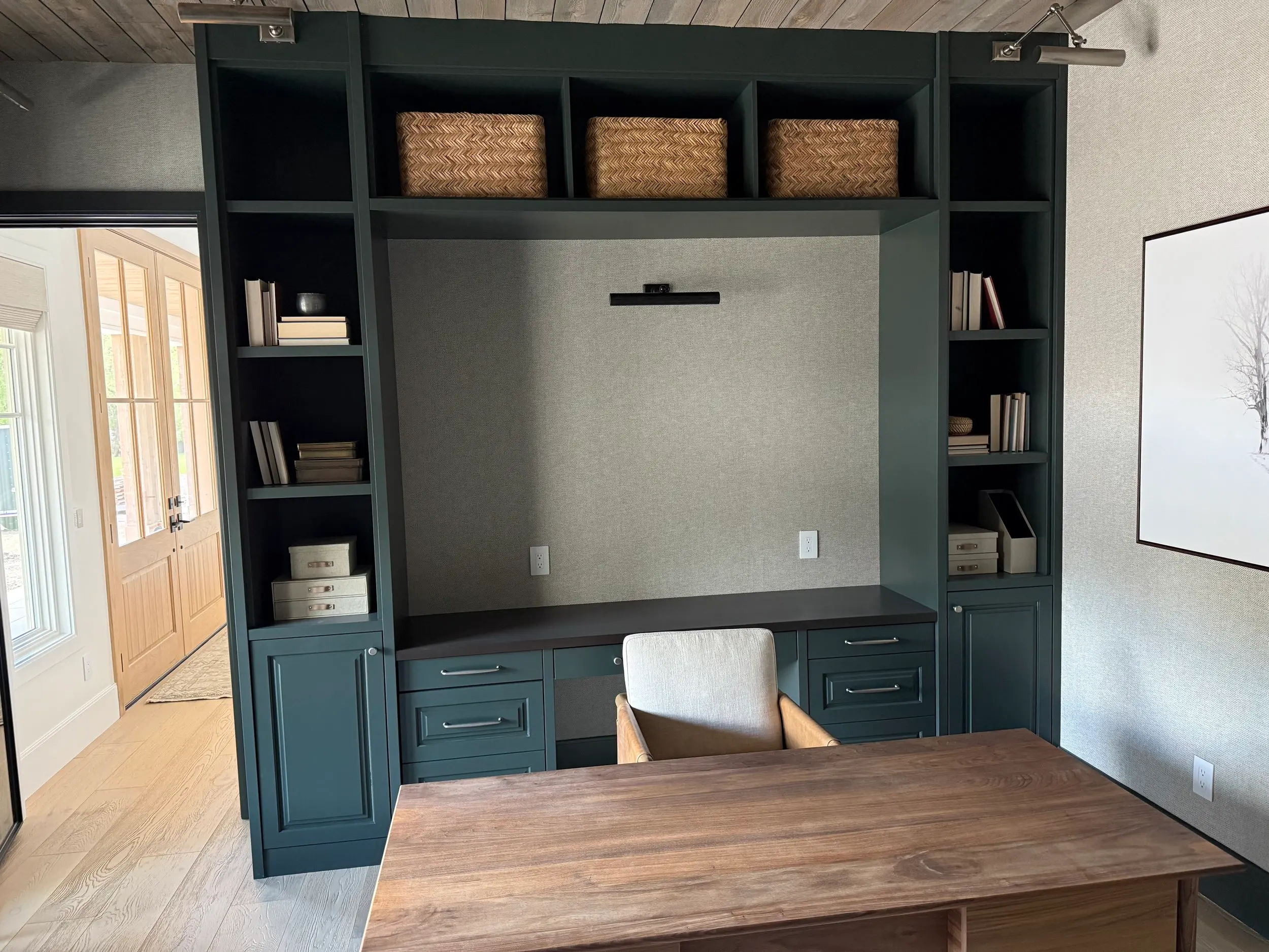 Dark green built-in home office desk with upholstered picture niche, woven storage baskets, and open bookshelves under a wood plank ceiling.