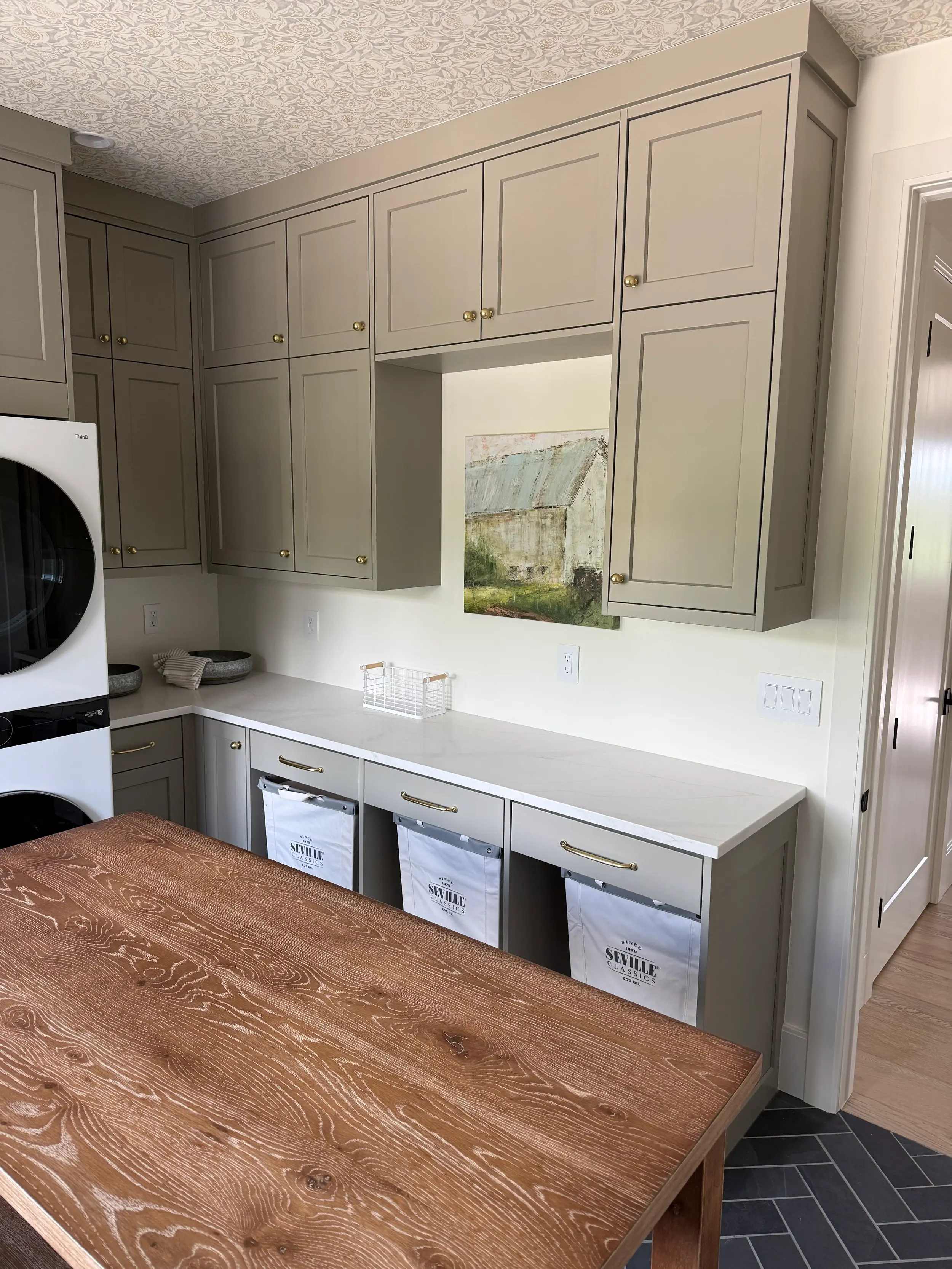 Taupe shaker laundry room cabinetry with quartz countertop, brass hardware, stacked washer-dryer, and canvas hamper bins on herringbone tile.